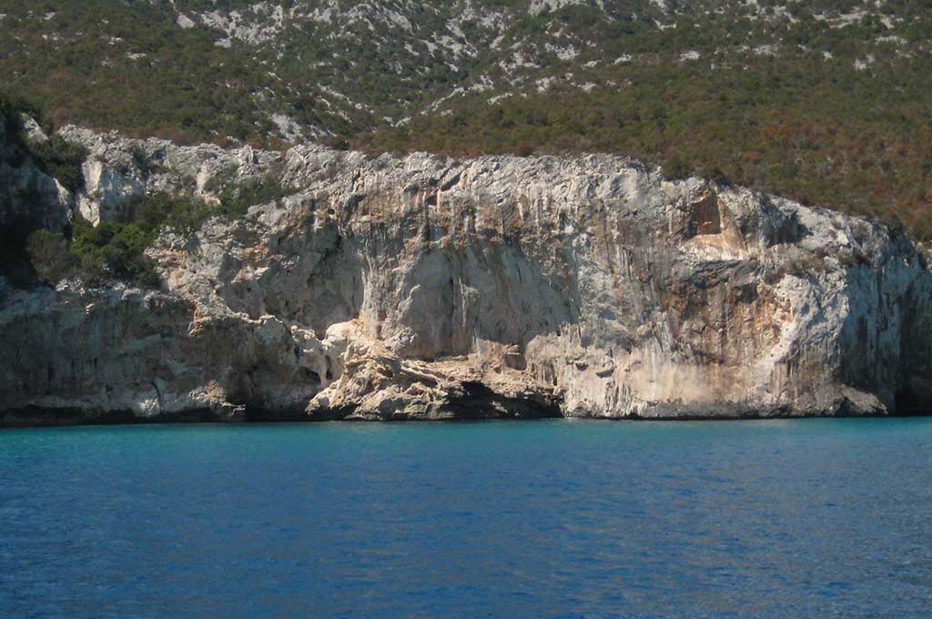 Das Bild zeigt ein einen Felsen über dem Meer bei Cala Gonone in Sardinien. Über dem blauen Meer steht ein weißer Fels mit überhängen, darüber Wald.