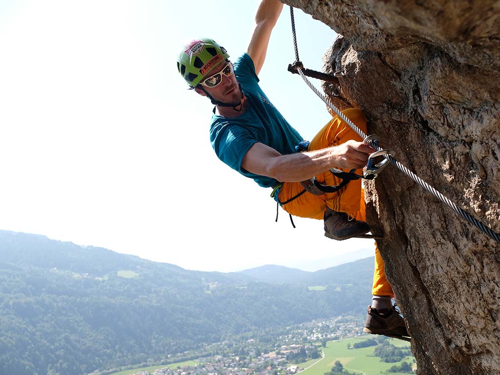 Das Bild zeigt einen Mann auf einem Klettersteig in großer Höhe. Er steht auf einem Klettersteig Trittbügel bzw. hält sich am Drahtseil fest und klippt sein Klettersteigset.