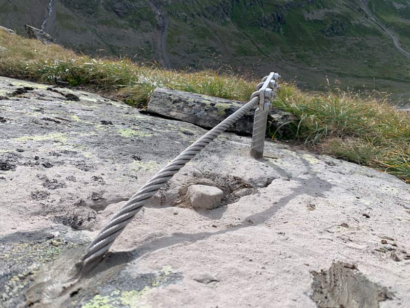 Das Bild zeigt den korrekten Verbau eines Klettersteig Anker. Ein Endanker ist im Hintergrund zu sehen aus welchem ein Drahtseil heraus kommt. Dieses ist im Vordergrund direkt im Fels eingeklebt.
