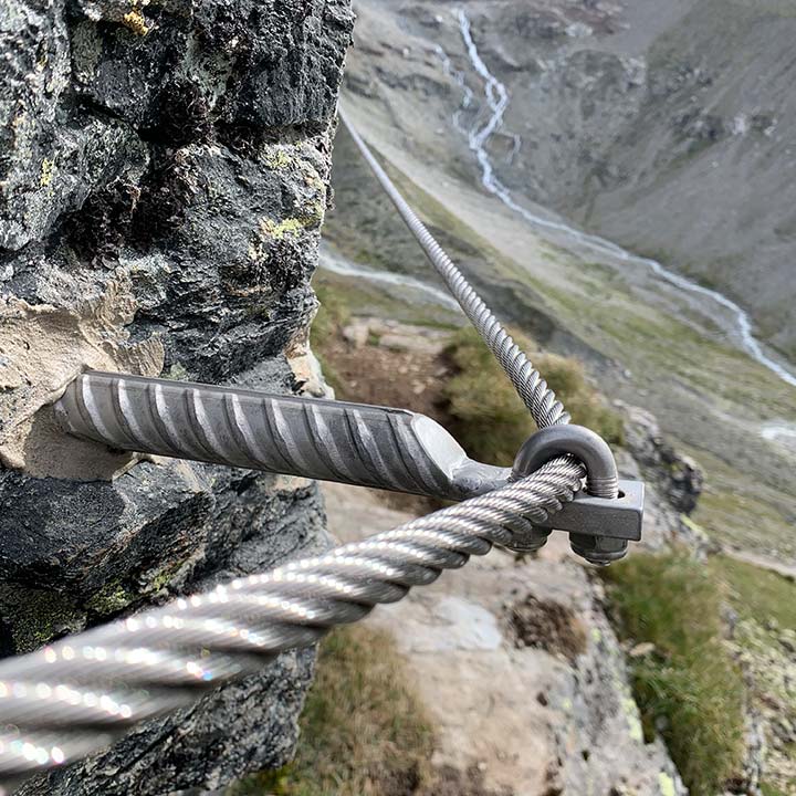 Das Bild zeigt ein Detail von einem Klettersteig. Ein Drahtseil läuft quer durch das Bild, es ist an einem Klemmanker befestigt. Im Hintergrund hochaline Landschaft.