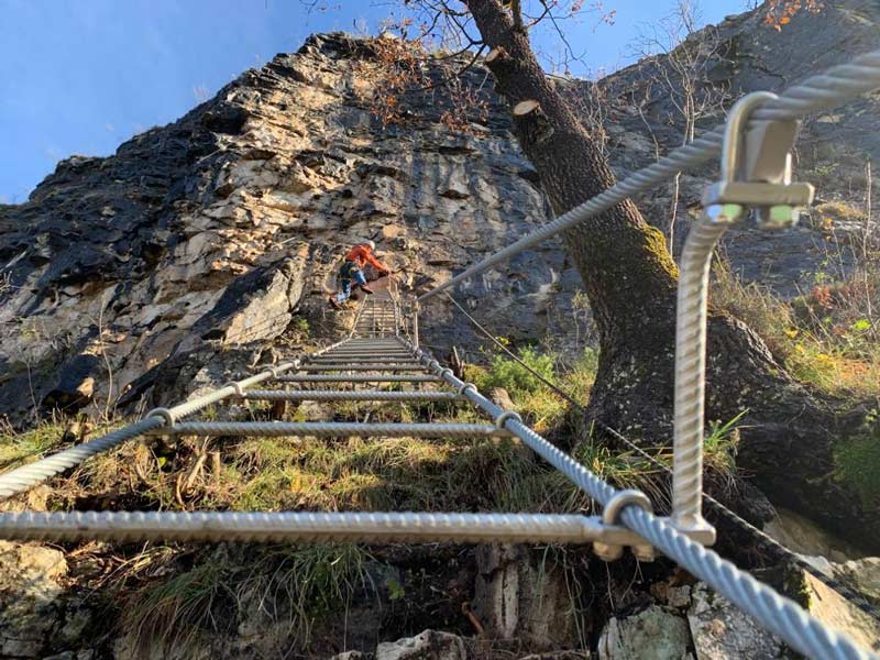 Das Bild zeigt die Verwendung einer Drahtseilstiegen Stufe beim Klettersteigbau. Die montierte Stufe ist im Vordergrund, im Hintergrund unscharf Fels, ein Kletterer und grüne Grasbüschel.