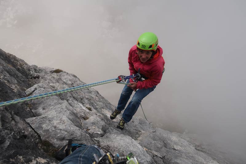 Das Bild zeigt eine Klettererin beim Abseilen mit Halbseilen in einer hohen Wand mit Nebel darunter.