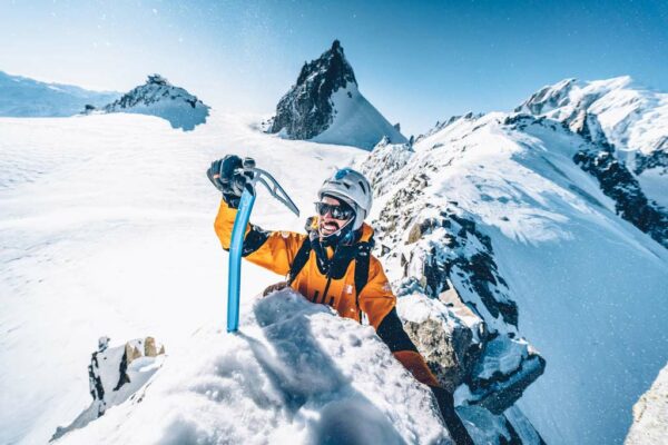 Das bild zeigt einen Alpinist an einem verschneiten Grat mit einem Eispickel im Schnee. Dahinter ein BErgpanorama.