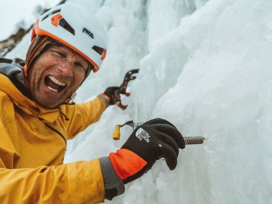 Das Bild zeigt einen Eiskletterer in oranger Jacke beim Eisschrauben setzen. Der Kletterer lacht in die Kamera.