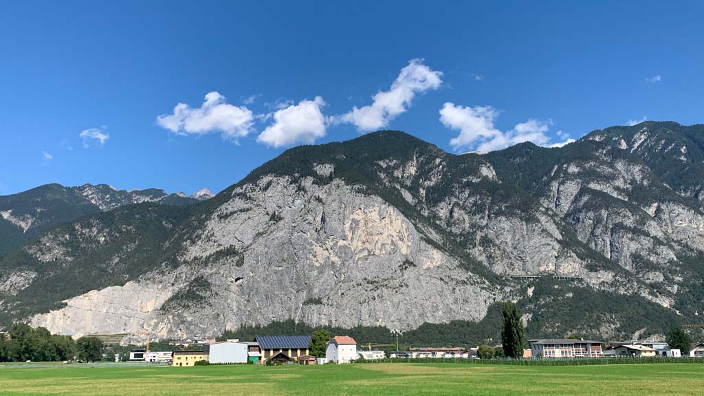 Das Bild zeigt die Martinswand bei Innsbruck. Hinter einem grünen Feld ragt eine hoher graue Felswand auf, darüber blauer Himmel.
