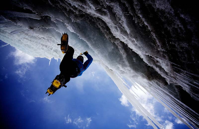 Das Bild zeigt einen Eiskletterer von unten in einem Eisvorhang und vor blauem Himmel.